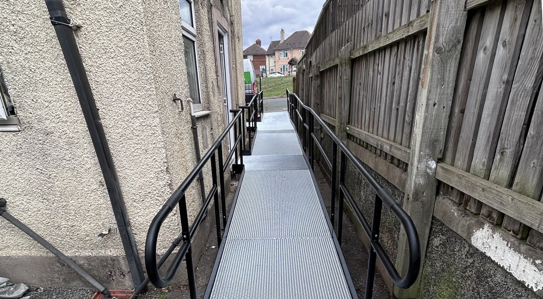 A metal modular wheelchair ramp with black handrails on both sides. The ramp runs alongside a house with a rough-textured wall on the left and a wooden fence on the right. It leads to an open space with houses visible in the background. The ramp has a grated surface for traction and is designed to provide safe access for people with mobility needs.