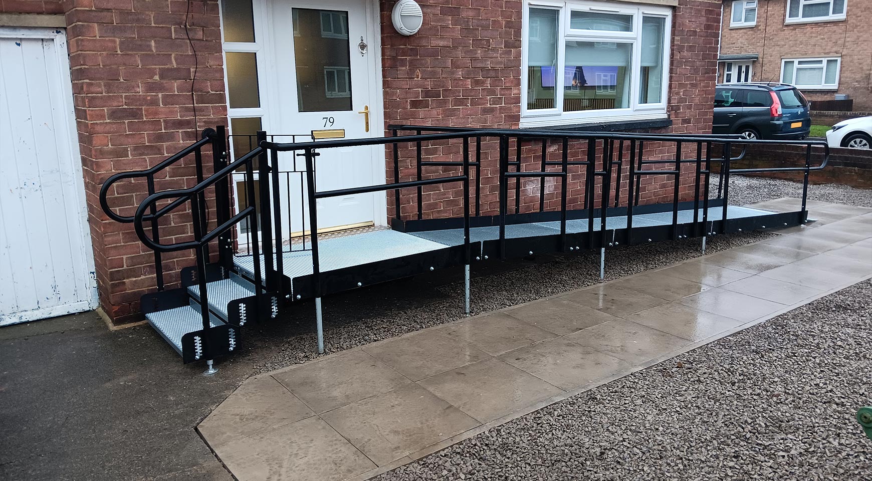 A Metal Modular Ramp with an attached step unit to the left. The ramp ascends from the right up to the door. This ramp is against a brick residential home with a white door.