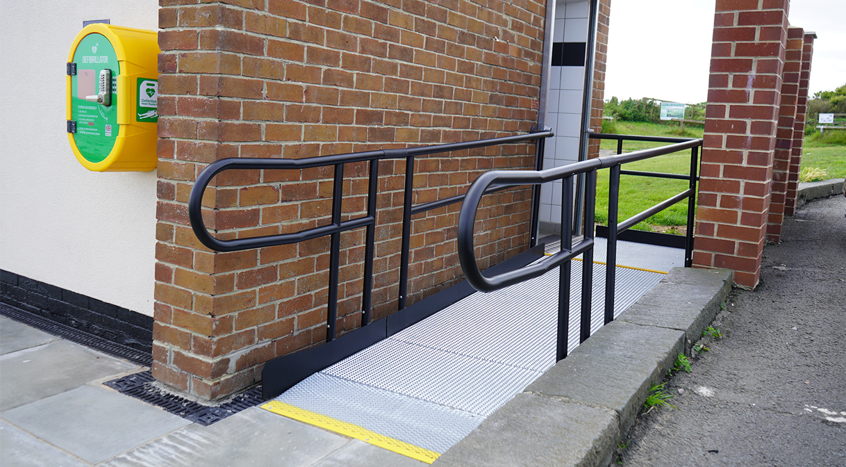 Black metal modular ramp that goes forward and turns left into a brick building. The ramp is right next to the brick wall. The surface of the ramp is a silver metal mesh.