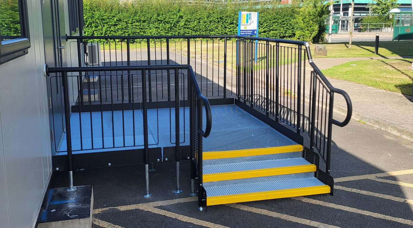 There is a omdular building to the left of the image that you can slightly see, and there is a modular step unit leading up to a platform which allows you to enter the door at the threshold height. The steps have yellow bull nosing on as a feature. The handrails of the steps and platform is black with a silver metal mesh platform.
