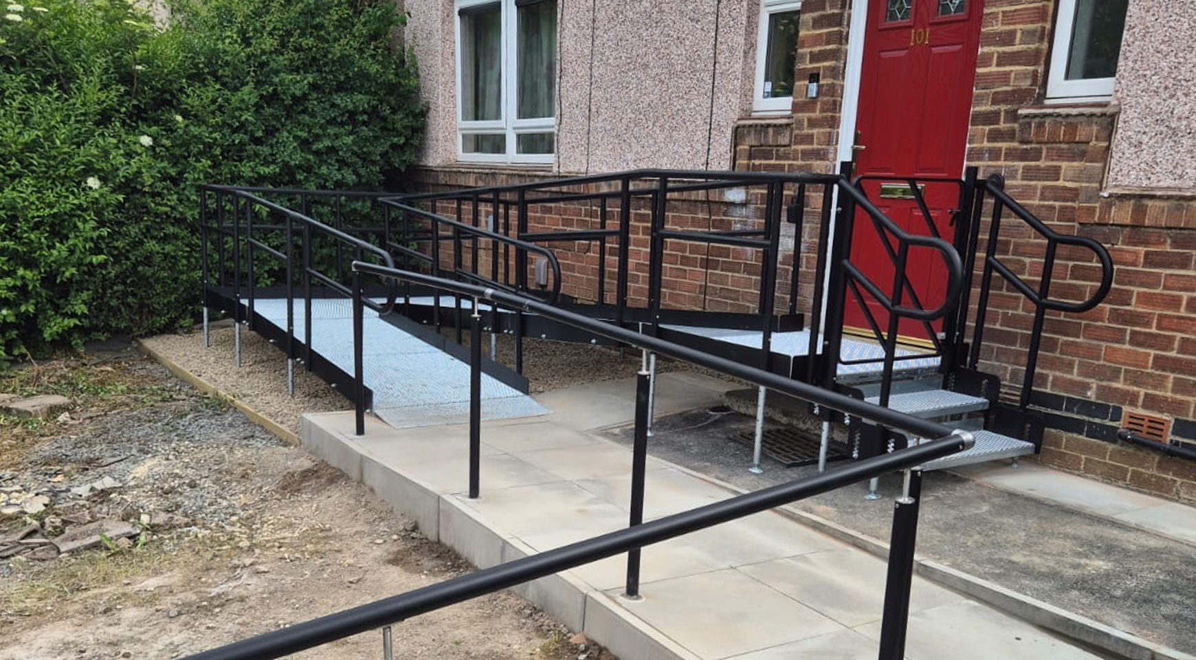 Image of a brick house with a red door. Infront of the door is a metal modular ramp that has black handrails and a silver metal mesh platform. The ramp goes parallel with the door, turns back on itself, which allows it to ramp to the front door. There is also a black handrail that starts just before the ramp and goes off down the driveway.